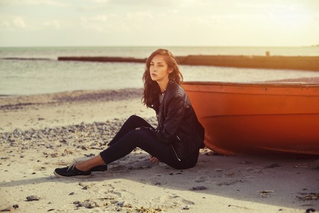 A girl sideways sitting near a red boat on the beach by the sea with beautiful sunset on the background. Woman in jeans, pullover and jacket on the sandの写真素材