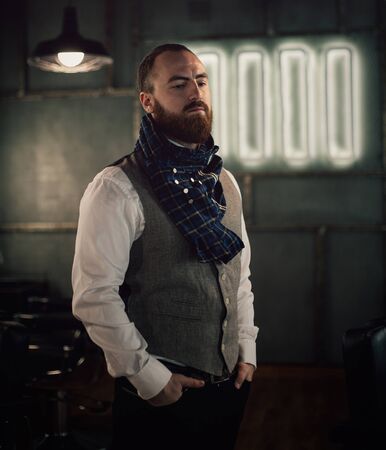 Handsome bearded man hipster with stylish beard in white shirt and chekered scarf standing with serious face in beauty salon or barbershopの写真素材