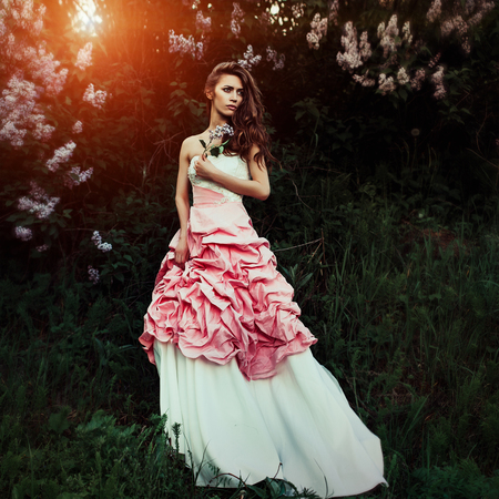 Beautiful and elegant woman with branch of lilacs and hair waves wearing wine red and white dress posing outdoors, retro vintage style and fashion. She looks like a runaway bride with dramatic emotionの写真素材