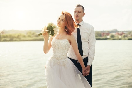 Elegant gentle stylish groom and bride near river or lake. Wedding couple in love. Woman in ehite dress man in white shirt and tie. With sunshine summer background.の写真素材