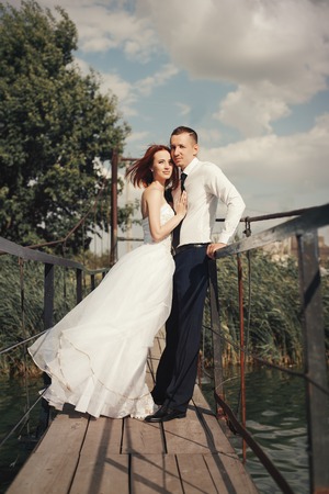 Wedding couple walking on bridge near lake on sunset at wedding day. Bride and groom in love. Woman in white dress main white shirt and tieの写真素材