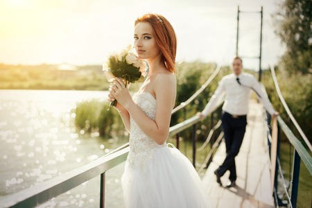redhead bride in white dress holding flower bouqet waiting for the groom in white shirt and tie standing on the bridge in the wedding summer dayの写真素材