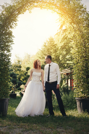 Handsome groom in white shirt and tie holds the bride's hand near green flower archway with beautiful sunset on the backgroundの写真素材