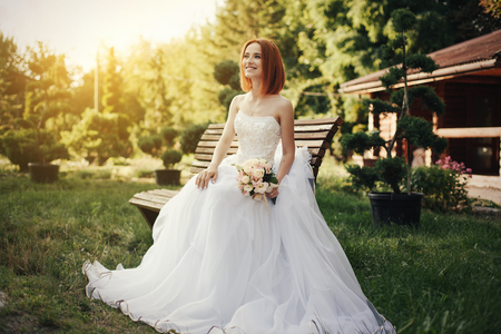 Bride in elegant white wedding dress gown sits on bench in the public park, with beautiful sunset on the backgroundの写真素材