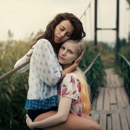 two beautiful young girlfriends blonde and brunette embracing on lake bridge on evening sunst. Rural scene. Women lesbians, hugging each otherの写真素材