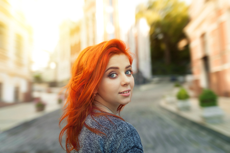 Startled young redhead woman looking back over her shoulder at the camera as she strolls down a deserted urban street. Girl with red hair and blue eyes dressed in sweeterの写真素材