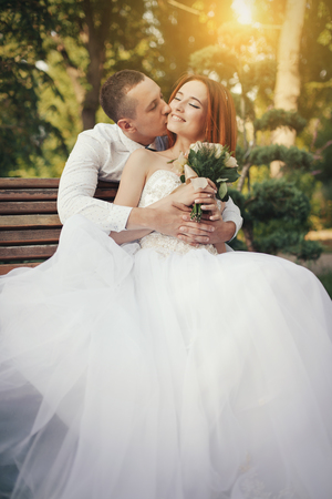 Bride in elegant white wedding dress with groom gown sits on bench in the public park, with beautiful sunset on the backgroundの写真素材