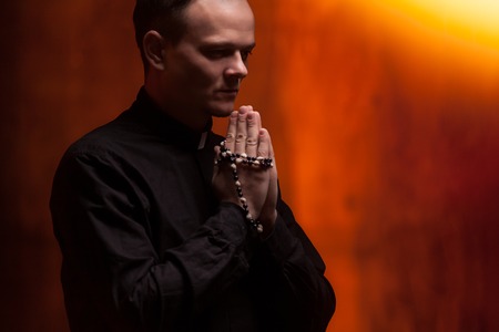 Portrait of handsome catholic priest or pastor with dog collar, dark red background. folded his hands in prayerの写真素材
