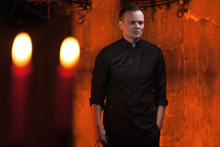 Portrait of handsome catholic priest or pastor with dog collar, dark red background. with candlesの写真素材