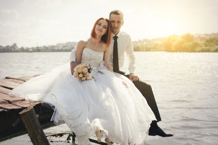 Wedding couple sitting on bridge near lake on sunset at wedding day. Bride and groom in love. Woman in white dress main white shirt and tieの写真素材