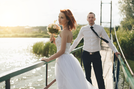Redhead bride in white dress holding flower bouquet waiting for the groom in white shirt and tie standing on the bridge in the wedding summer dayの写真素材