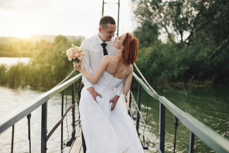 Redhead bride in white dress holding flower bouquet and groom in white shirt and tie are standing on the bridge in the wedding summer dayの写真素材