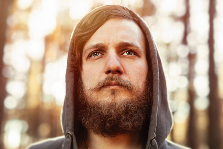 close up portrait of a bearded hipster tourist in gray hood man in the sunlight woods forest