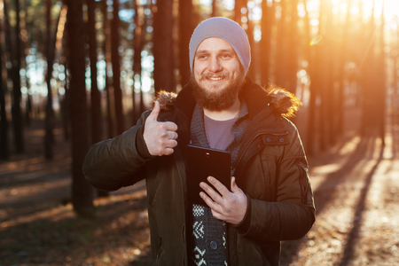 portrait of smiling a bearded hipster man in the woods forest with thumbs up and black tablet in handの写真素材
