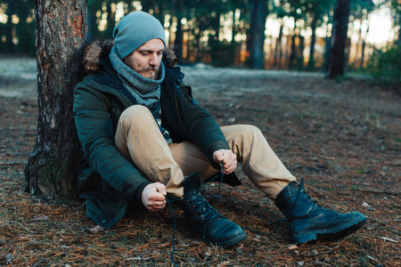 bearded hipster tourist man in the woods forest sitting near a tree tying shoelaces restingの写真素材