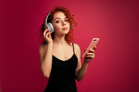 beautiful redhead girl in big white headphones with phone in hand listening to music in the Studio on a pink backgroundの写真素材
