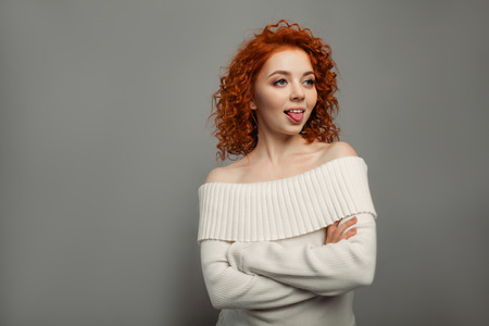 young cute redhead curly haired girl shows her tongue teasing on the gray backgroun in studioの写真素材