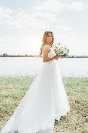 Bride in a white dress standing in a green field near the river and holding a wedding bouquet of flowers and greeneryの写真素材