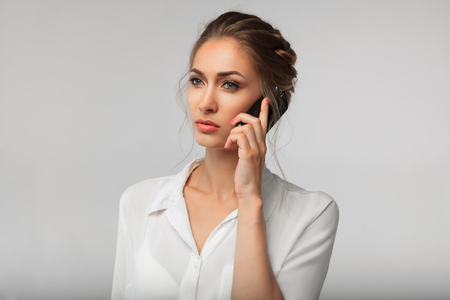 beautiful businesswoman dressed in white office shirt with a cell phone in hands. serious conversation. business portrait on the gray backgroundの写真素材