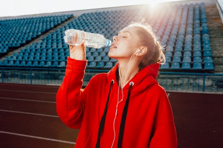 People, lifestyle and sport concept. Sport woman athlet overcame long distance, drinks water from plastic bottle, listens audio tracks in earphones. Dressed in the urban casual clothes. Bright red hoodieの写真素材