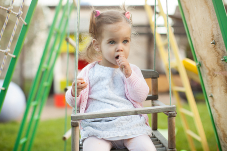 Funny little child, adorable preschooler girl in pretty dress having fun on a swing in the park on summer day With food in her handの写真素材