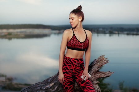 Healthy lifestyle and sport concepts. Woman in fashionable sportswear rests after a sports active training near the lake. Listens to music on headphones with a smartphone in hands Evening timeの写真素材