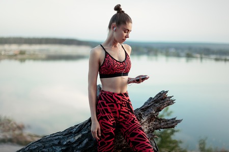 A beautiful brunette girl in stylish sports clothes is resting after a sport active workout near the lake. Listens to music on headphones with a smartphone in hands Healthy lifestyle and sport concepts.の写真素材