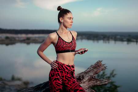 A beautiful brunette girl in stylish sports clothes is resting after a sport active workout near the lake. Listens to music on headphones with a smartphone in hands Healthy lifestyle and sport concepts.の写真素材