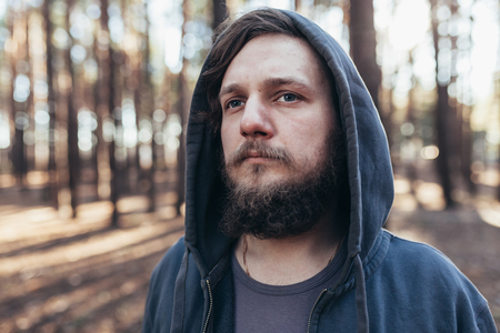a young man with a beard walks in a pine forest. Portrait of a brutal bearded man in a hood Homelessの写真素材