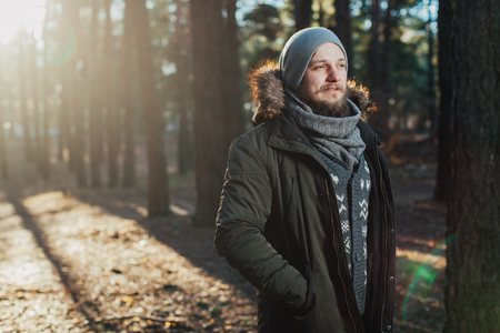 Portrait, close-up of young stylishly serious man with a beard dressed in rgreen winter jacket with a hood and fur on his head stands against the background of pine forestの写真素材