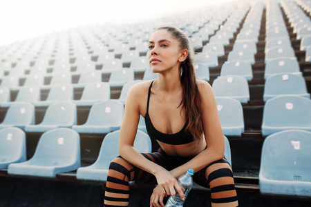 Sporty young attractive girl in sportswear relaxing after hard workout sit and drink water from bottle after running on a stadium Summer workoutの写真素材