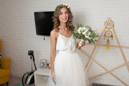 The bride in a beautiful wedding dress with a long curly hair stands in the bedroom and looks at the camera. Bride indoors with white flowers bouquet and tender wreath hairstyle.の写真素材