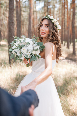 Follow me my love concept. Attractive young woman dressed in white wedding dress holding hand of her boyfriend and walking on the beautiful garden to happy future.の写真素材
