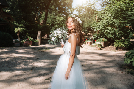 A cute curly woman in a white wedding dress with a wedding bouquet and wreath in her hair standing back to the camera in nature. Concept escaped bride. Forward to a happy bright future Runaway.の写真素材