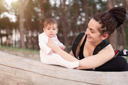 A beautiful smiling young brunette woman with long dreadlock hair hold a pretty baby. Mother looks tenderly at the child. They are in the green summer forest. With sunshine on the backgroundの写真素材