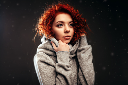 young cute redhair girl with blue eyes wrapped himself in a gray hoodie large portrait in studio on the black backgroundの写真素材