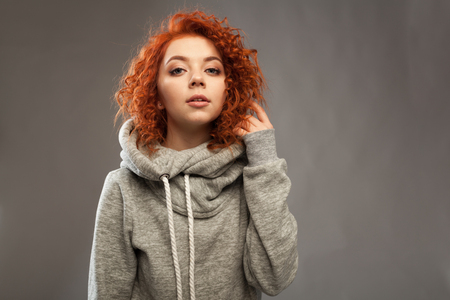 Portrait of a beautiful young curly-haired girl with fiery red hair looking at the camera on a gray background. Studio photography. Warm clothes.の写真素材