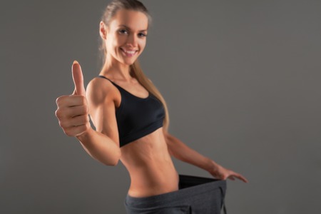 Slim young woman wearing old big pants, showing successful result of a diet - weight loss, slimming concept. gray background Happy wellnes lifeの写真素材