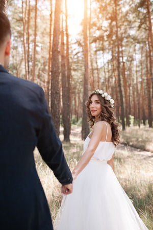 Follow me my love concept. Attractive young woman dressed in white wedding dress holding hand of her boyfriend and walking on the beautiful garden to happy future.の写真素材