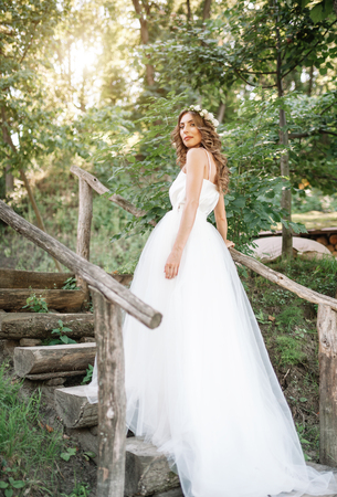 A cute curly woman in a white wedding dress with a wedding bouquet and wreath in her hair standing back to the camera in nature. Concept escaped bride. Forward to a happy bright future Runaway.の写真素材