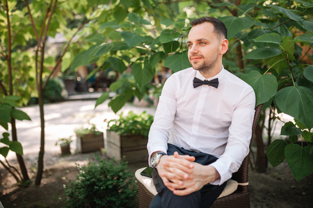 A handsome young business man in a white shirt and tie a butterfly at a business dinner waits for his colleagues alone at a summer restaurant table.の写真素材