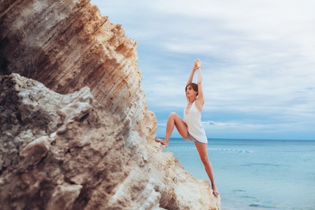A beautiful young girl with short hair is dressed in shorts and a white jersey practicing yoga against the backdrop of the sea and mountains. Pose warrior hero. Concept about balance and calmness.の写真素材