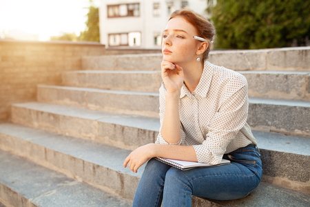 beautiful young redhead girl with freckles sitting on a stairs and making notes in a notebook. Portrait of a student. Remote education. Homework.の写真素材
