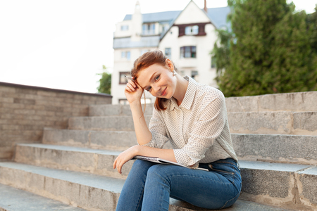 beautiful young redhead girl with freckles sitting on a stairs and making notes in a notebook. Portrait of a student. Remote education. Homework.の写真素材