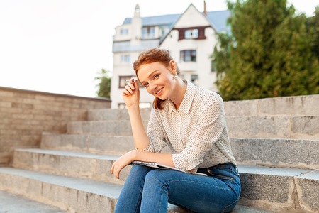 Young beautiful red hair girl with freckles sitting on a stairs near the university writing in a notebook with homework. Portrait of a student. Back to school conceptの写真素材