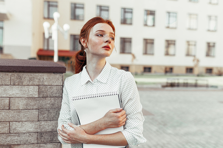 Beautiful red haired girl with freckles hugging notebooks with homework and smiling happy. Back to school concept. Portrait of a student.の写真素材