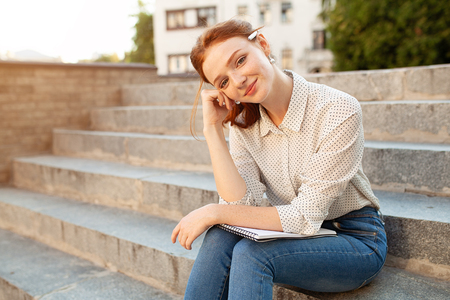 A beautiful young redhaired girl writes a romantic love letter sitting on a stairs near the institute. Student love. Or homework outdoorsの写真素材