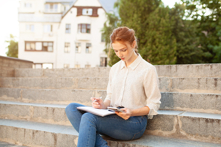 Young beautiful red hair girl with freckles sitting on a stairs near the university writing in a notebook with homework. Portrait of a student. Back to school conceptの写真素材