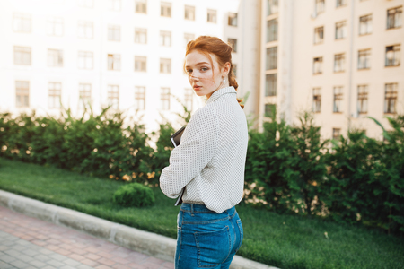 Rear view of a teenager student red hair girl with fresckles walking on a path at a college campus grounds carrying a notepad and turning to smile at the camera joyfully. Student lifestyle outdoorsの写真素材