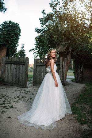 A cute curly woman in a white wedding dress with a wedding bouquet and wreath in her hair standing back to the camera in nature. Concept escaped bride. Forward to a happy bright future Runaway.の写真素材